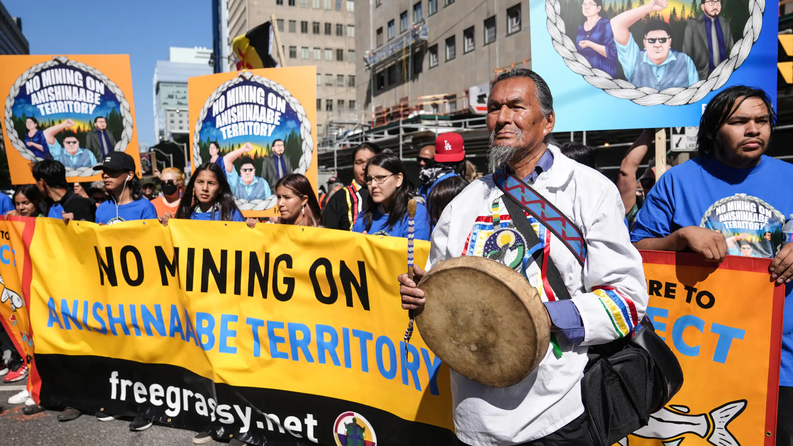 Un groupe d'autochtones brandit des bannières avec des slogans s'opposant aux mines, et l'un deux, à l'avant-plan, tient un tambour traditionnel.