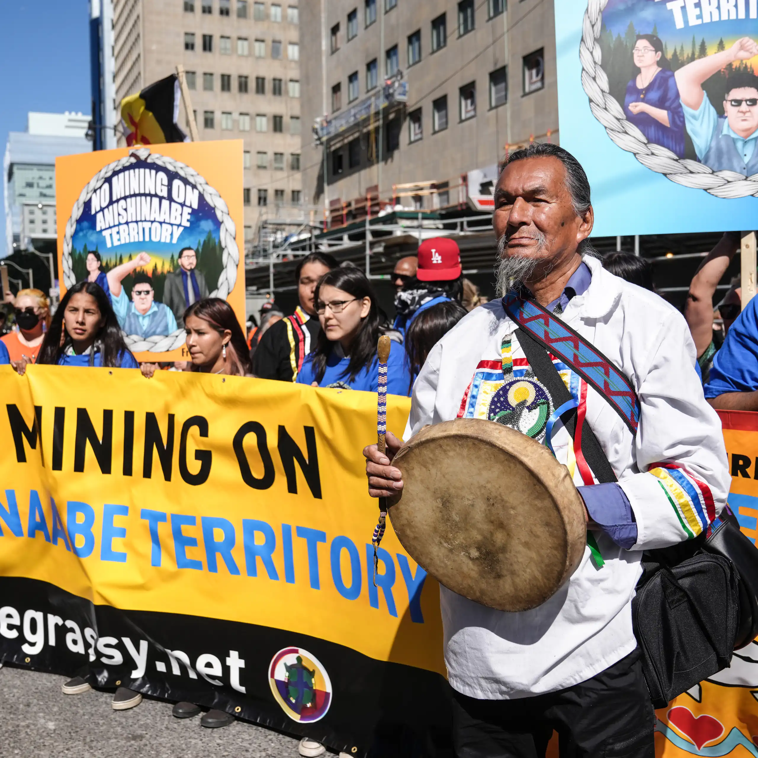 Un groupe d'autochtones brandit des bannières avec des slogans s'opposant aux mines, et l'un deux, à l'avant-plan, tient un tambour traditionnel.