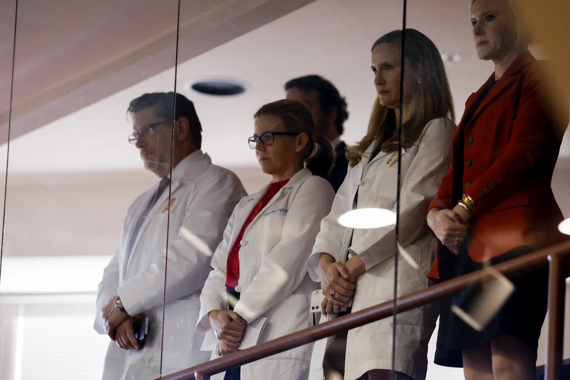 Five people, three of whom wear white doctor coats, stand behind a pane of glass, holding their hands in front of them.