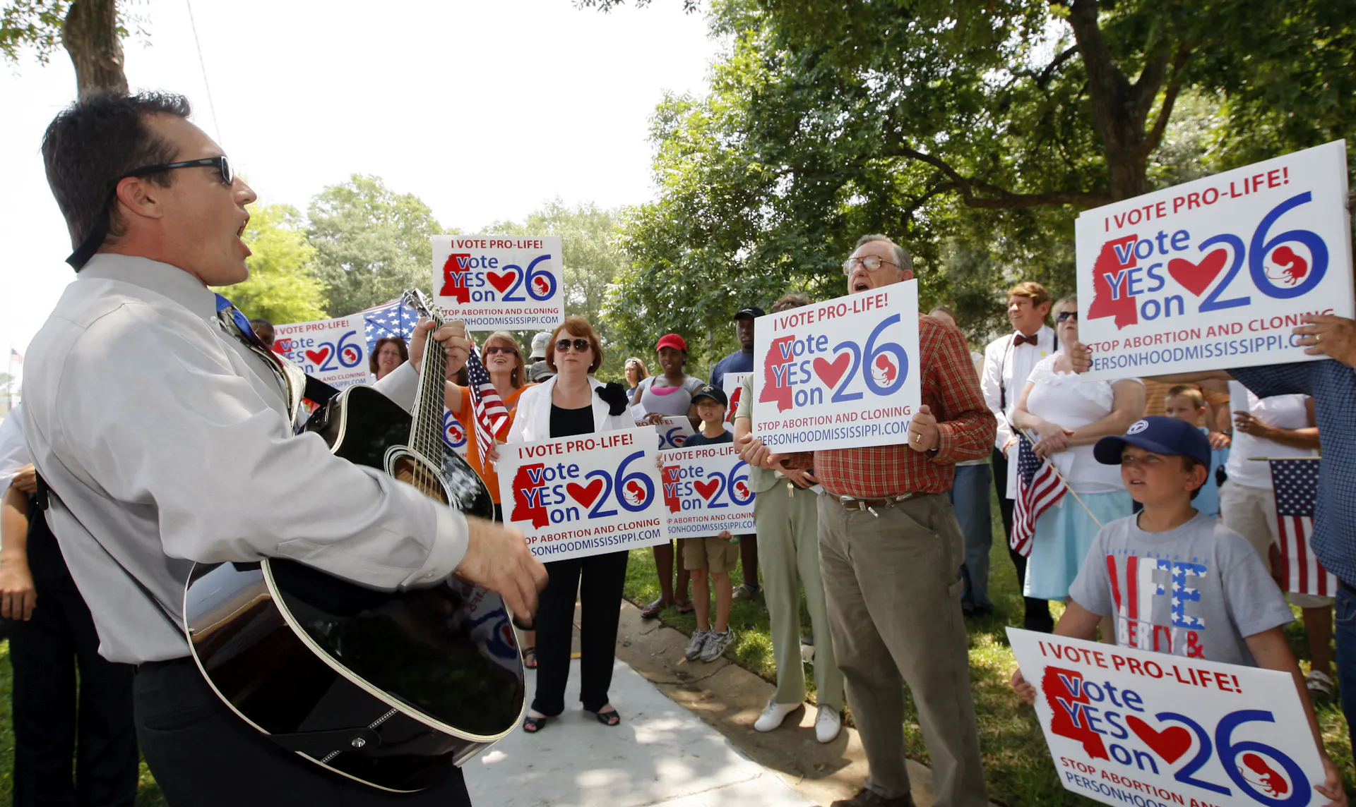 A man plays a guitar to a crowd of people outside holding signs that say 'I vote pro-life!'