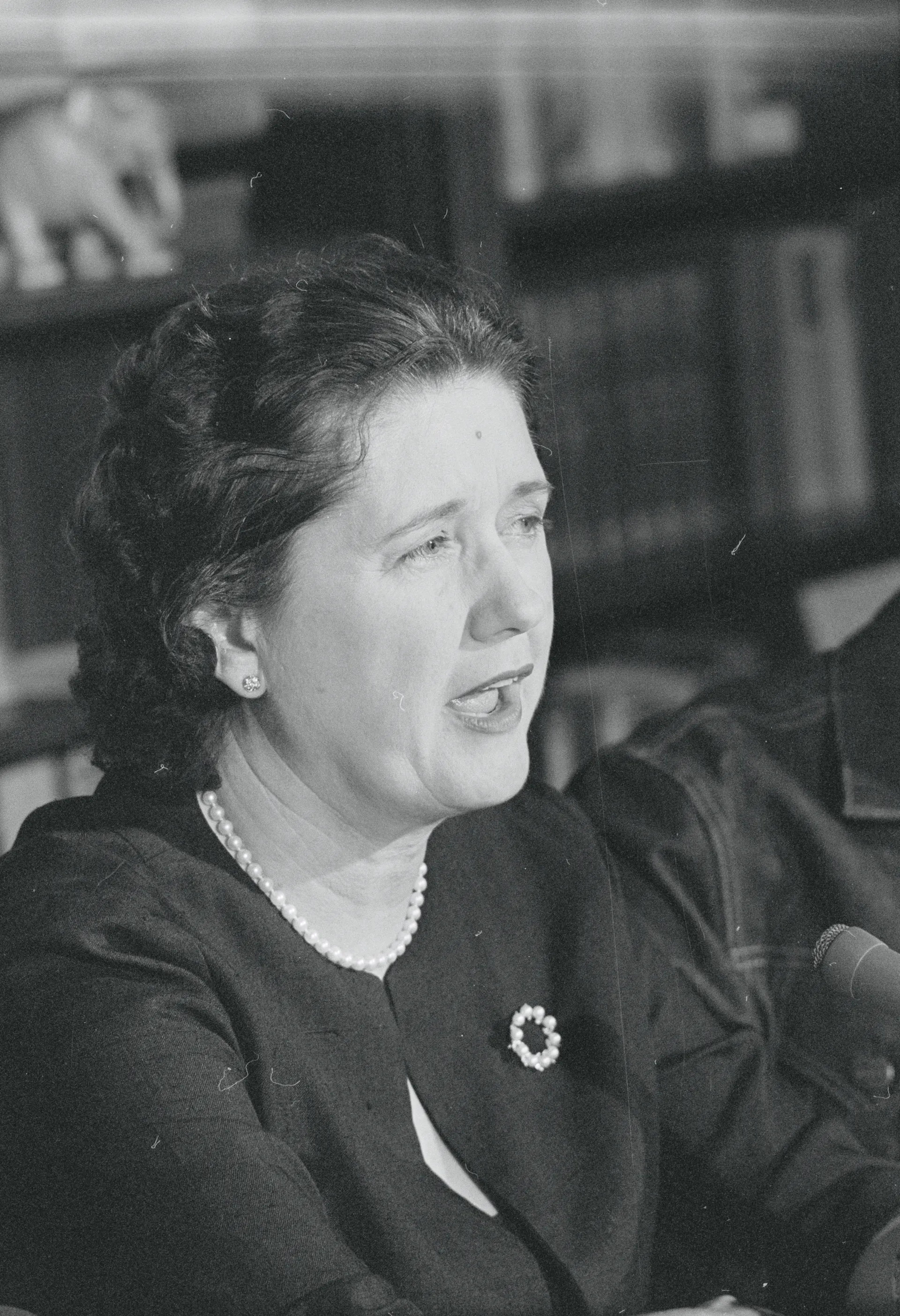 A dark-haired woman wearing a strand of pearls and a dark top, speaking into a microphone.