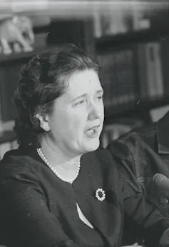 A dark-haired woman wearing a strand of pearls and a dark top, speaking into a microphone.