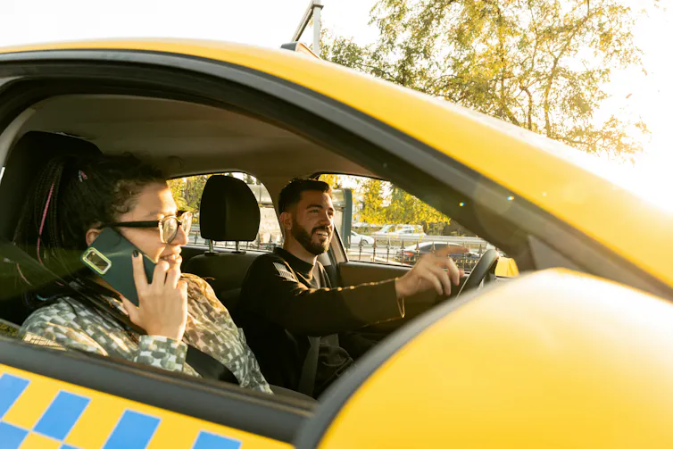A taxi driver smiles in the car next to a passenger, who is on the phone
