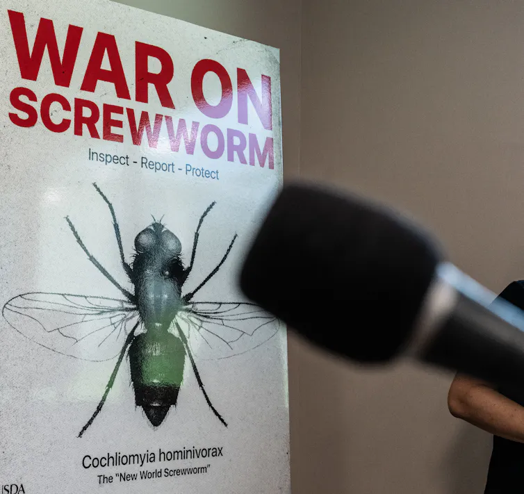 A microphone is placed in front of a white sign with the words war on screwworm in red lettering and a picture of a green fly