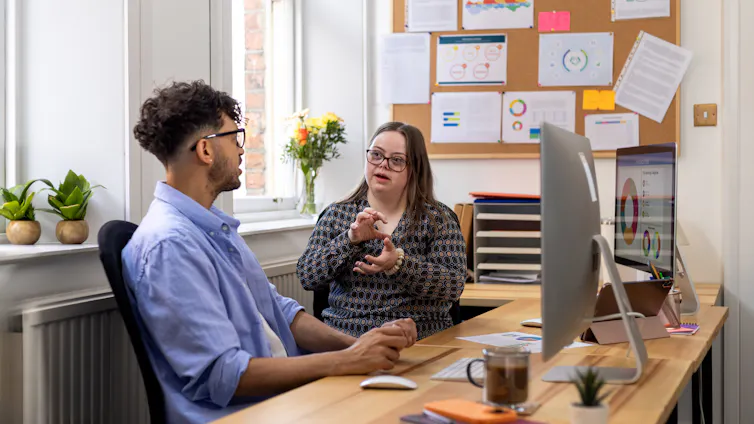 Medium shot of a young adult female with Down syndrome and her mid-adult male colleague sitting at a desk in an office