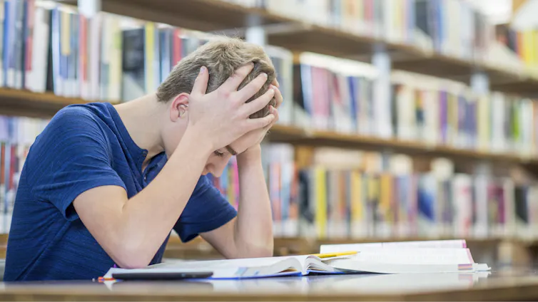 A teenage boy appears stressed as he holds his head in his hands while reading a book in the library.