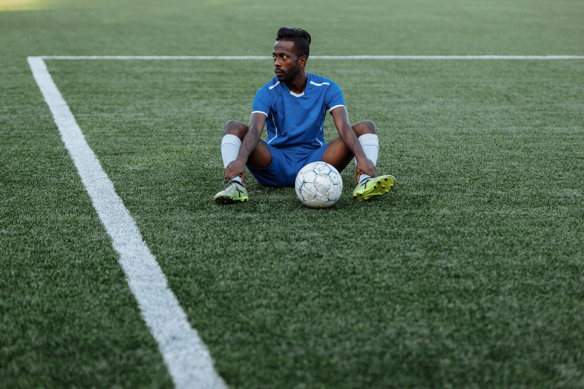 A young African football player sits on a marked field with a soccer ball between his legs. He looks off in the distance, dejected and defiant.