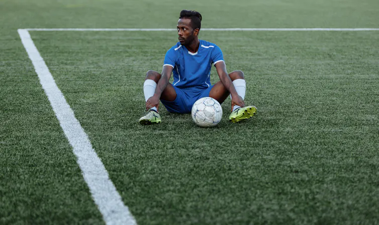 A young African football player sits on a marked field with a soccer ball between his legs. He looks off in the distance, dejected and defiant.