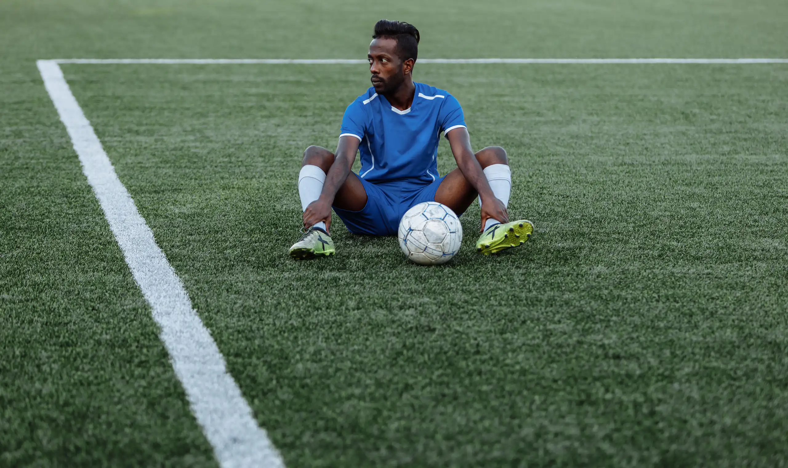 A young African football player sits on a marked field with a soccer ball between his legs. He looks off in the distance, dejected and defiant.