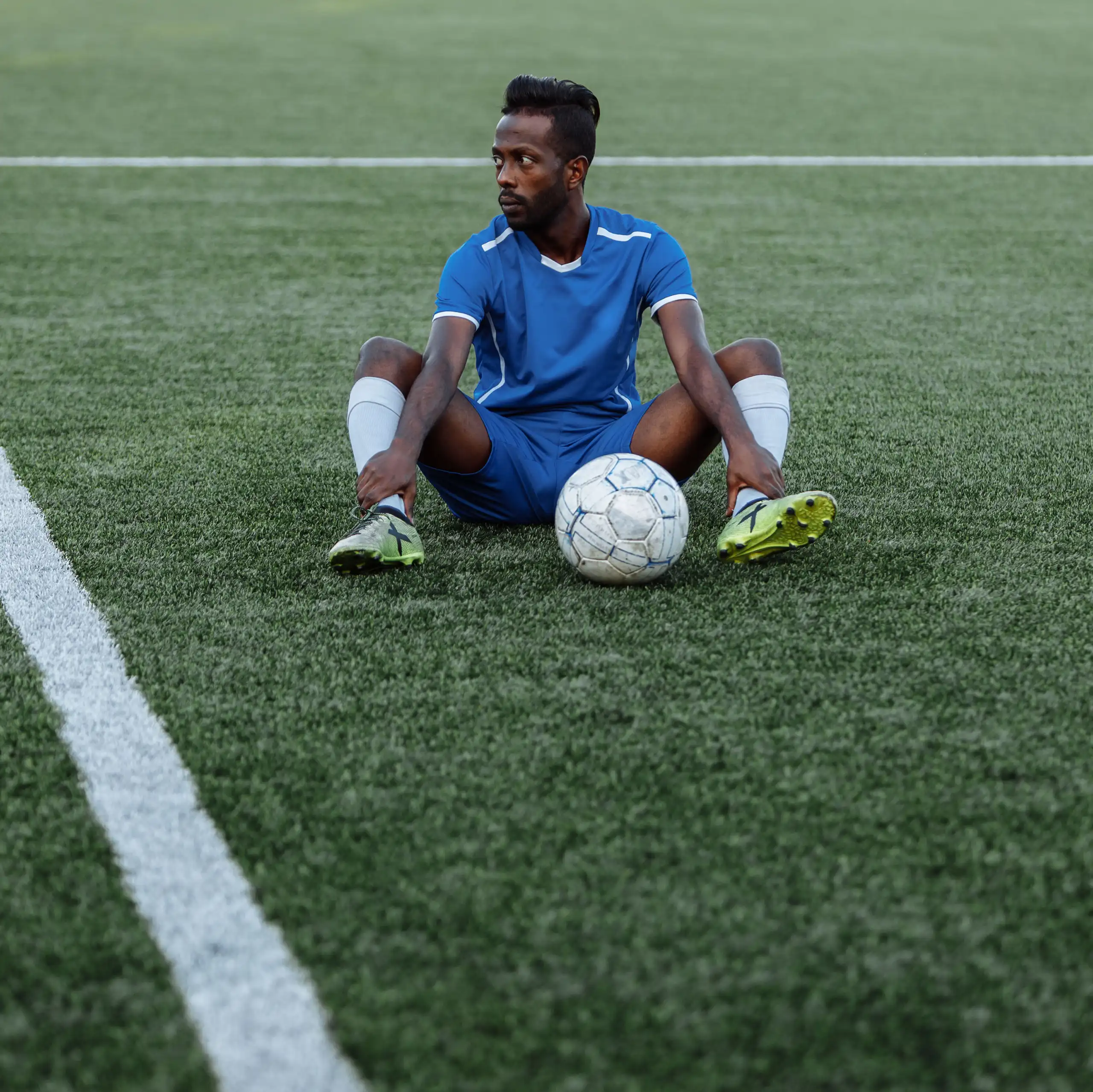 A young African football player sits on a marked field with a soccer ball between his legs. He looks off in the distance, dejected and defiant.