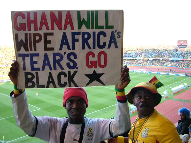 Two African men on a sports stand, one holding up a hand painted poster that reads: 'Ghana will wipe Africa's tears. Go Black (and a star).'