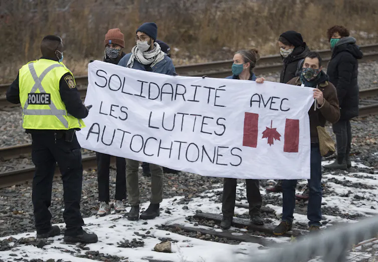 Un policier parlemente avec des manifestants qui brandissent en travers de la voie ferrée une bannière avec l’inscription « Solidarité avec les luttes autochtones. »
