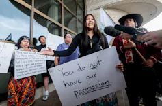 A young woman holding a placard, surrounded by other protesters, speaks into microphones from various media outlets
