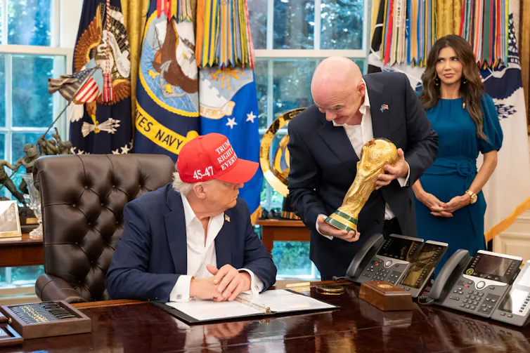 A fat old guy in a red baseball hat and a short bald guy holding the World Cup trophy in the Oval Office.