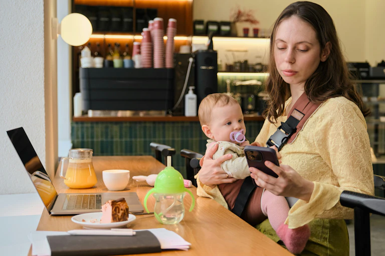 A mother with a baby in a carrier trying to work at a laptop.