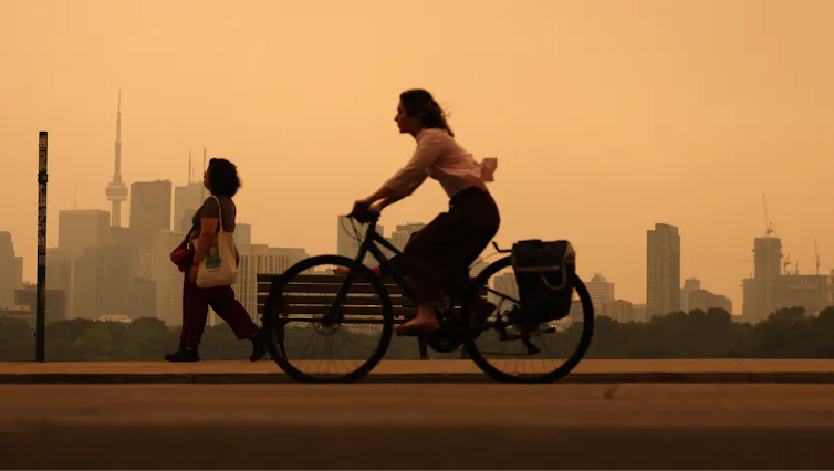 A woman rides a bike through city air tinted orange by wildfire smoke. Neither she nor a woman walking in the background is wearing a protective mask.