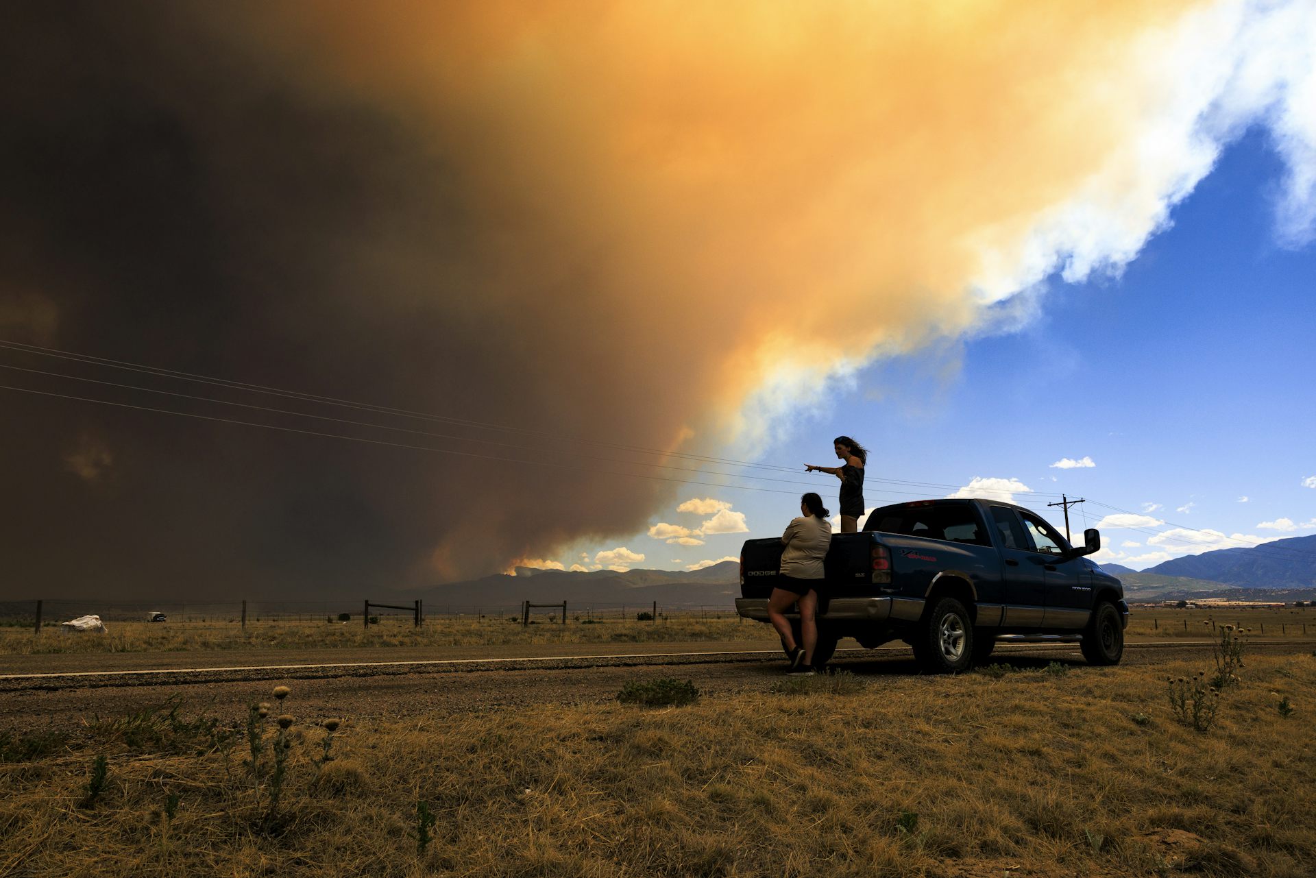 La gente permanece parada en un campo mientras una columna de humo tiñe el cielo de naranja y luego se oscurece.