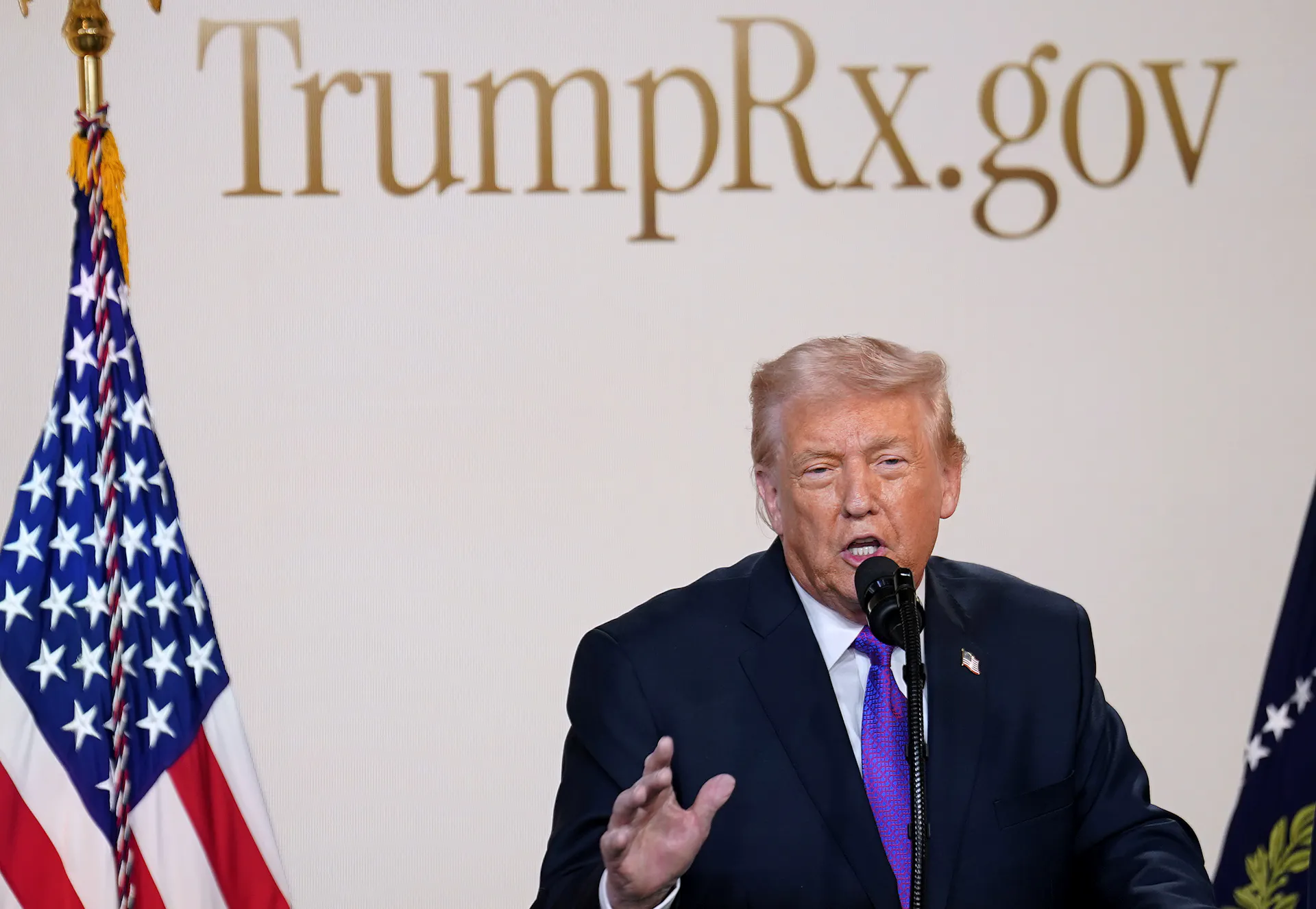 A man with a purple tie speaks behind the podium with the word TrumpRx.gov behind him in gold lettering and an American flag to his right.