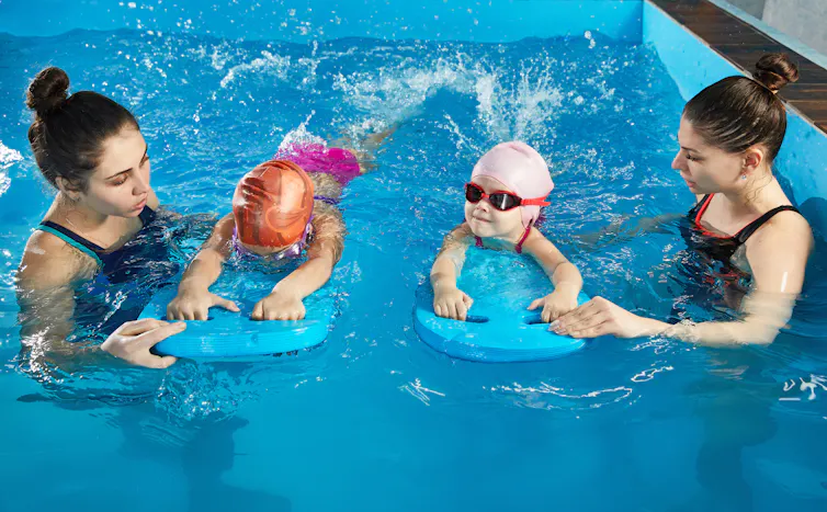 Two young children holding paddleboards work one-on-one with instructors in the pool.