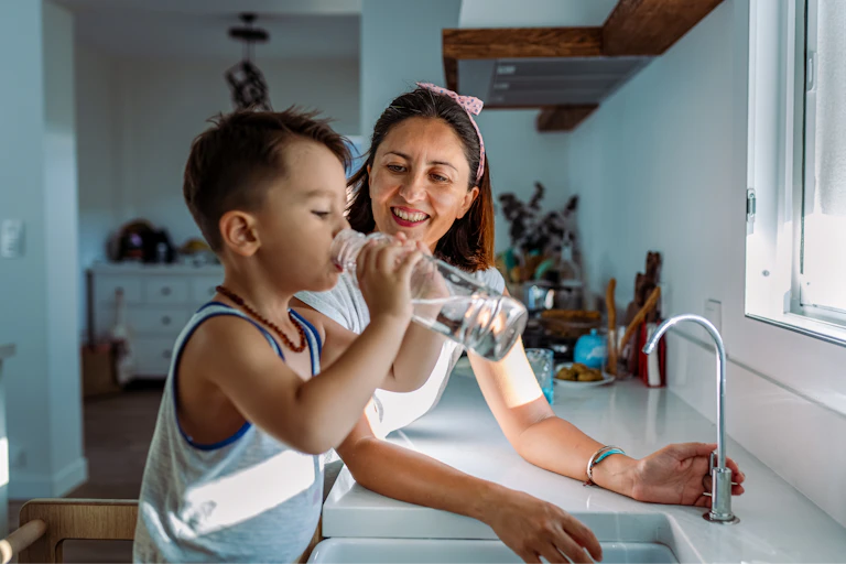 Mother and young boy stand by sink as boy drinks bottle of water