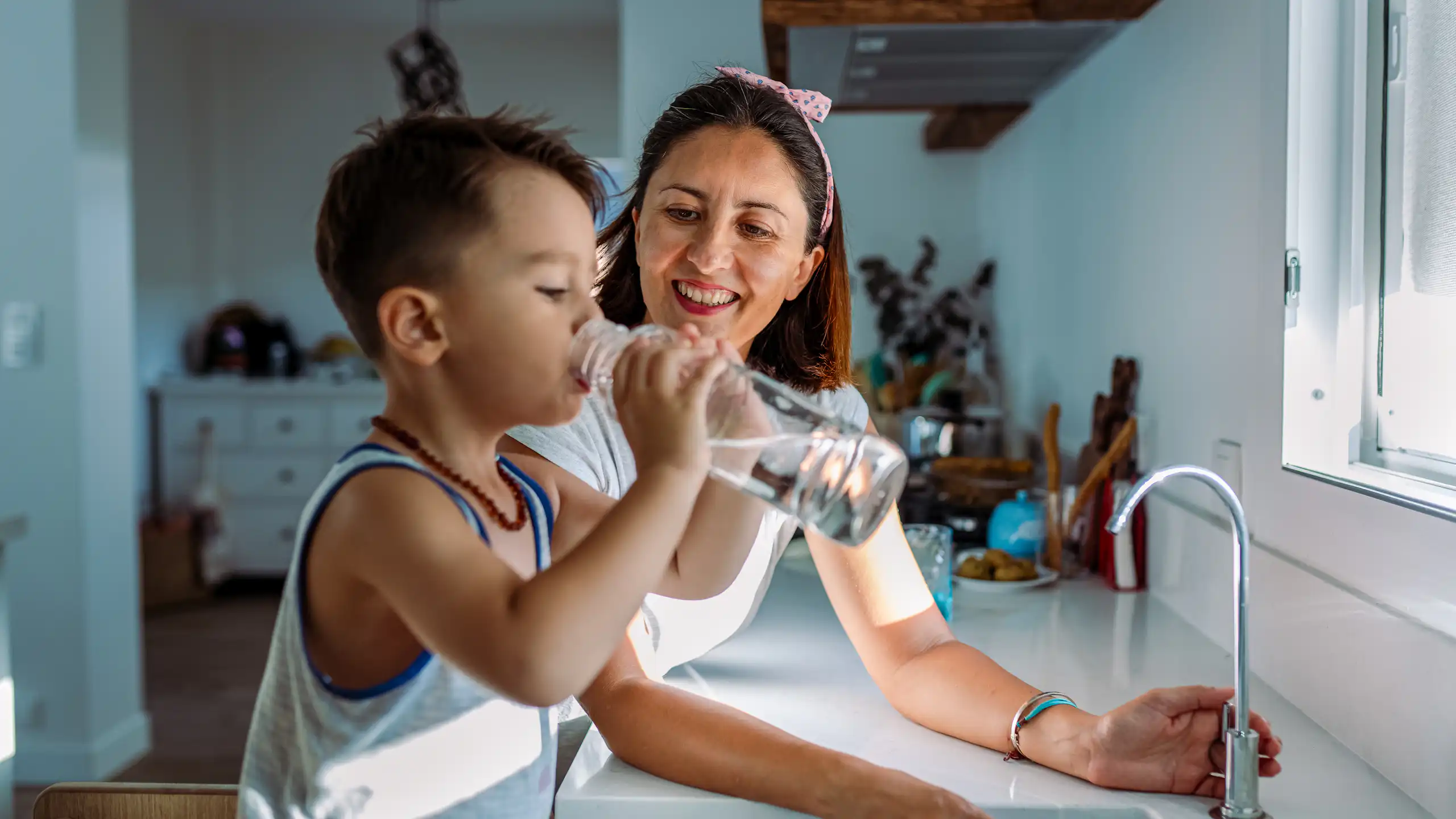 Mother and young boy stand by sink as boy drinks bottle of water