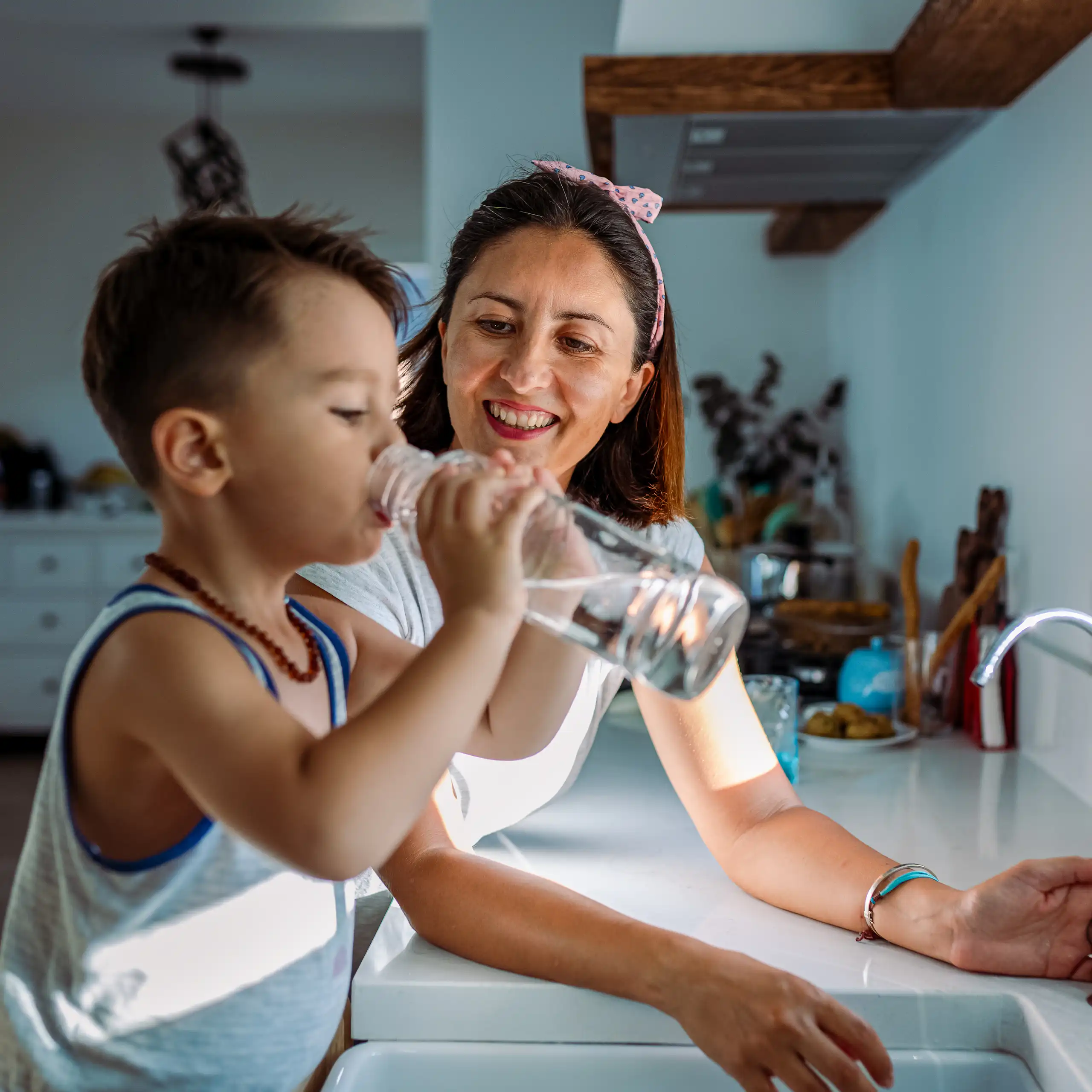 Mother and young boy stand by sink as boy drinks bottle of water