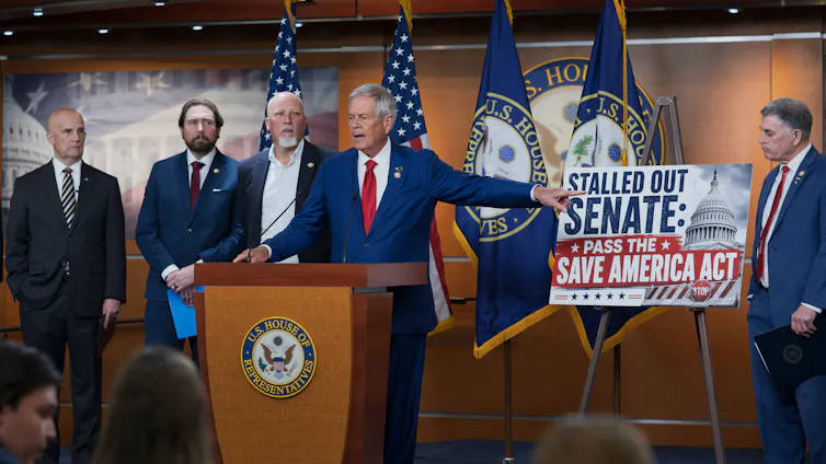 Stern men in suits stand behind a lectern, with one pointing to a sign chastising the U.S. Senate.