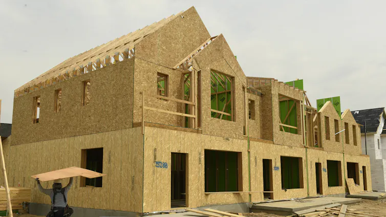 Tan panels with open cut-outs for windows create the frame for a housing structure, while wood planks are on the ground at the structure's base.
