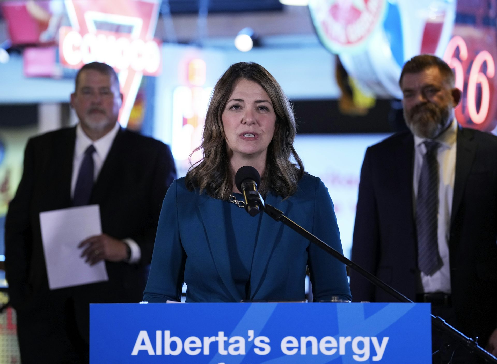 A middle-aged white woman with shoulder-length hair in a blue suit speaks from behind a podium