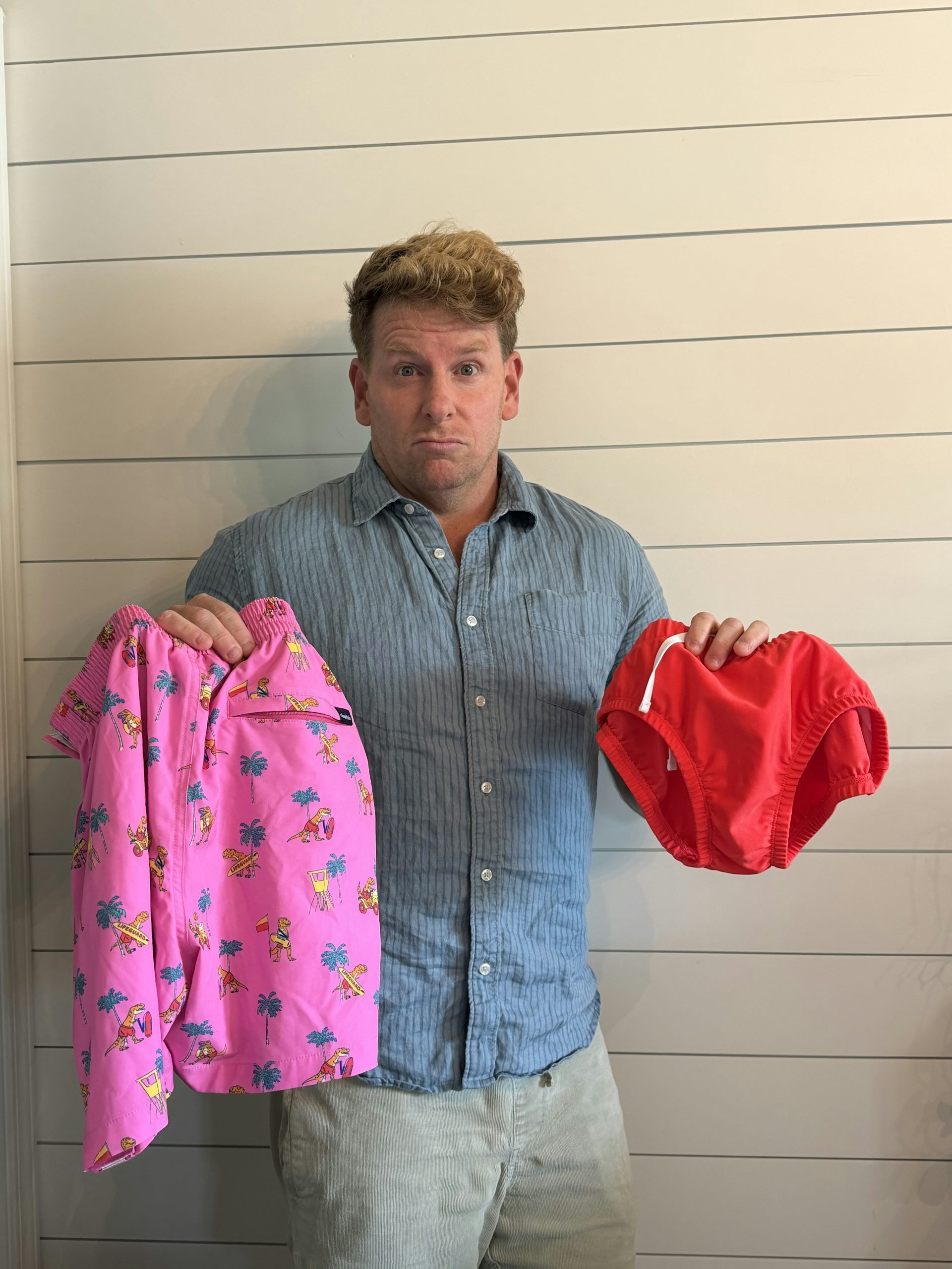 A man in a blue shirt holds up a pair of Red Speedos and a dinosaur-themed wrap.