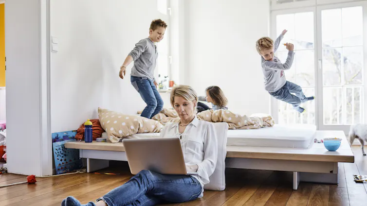 A woman works on a laptop while three kids jump on a bed.