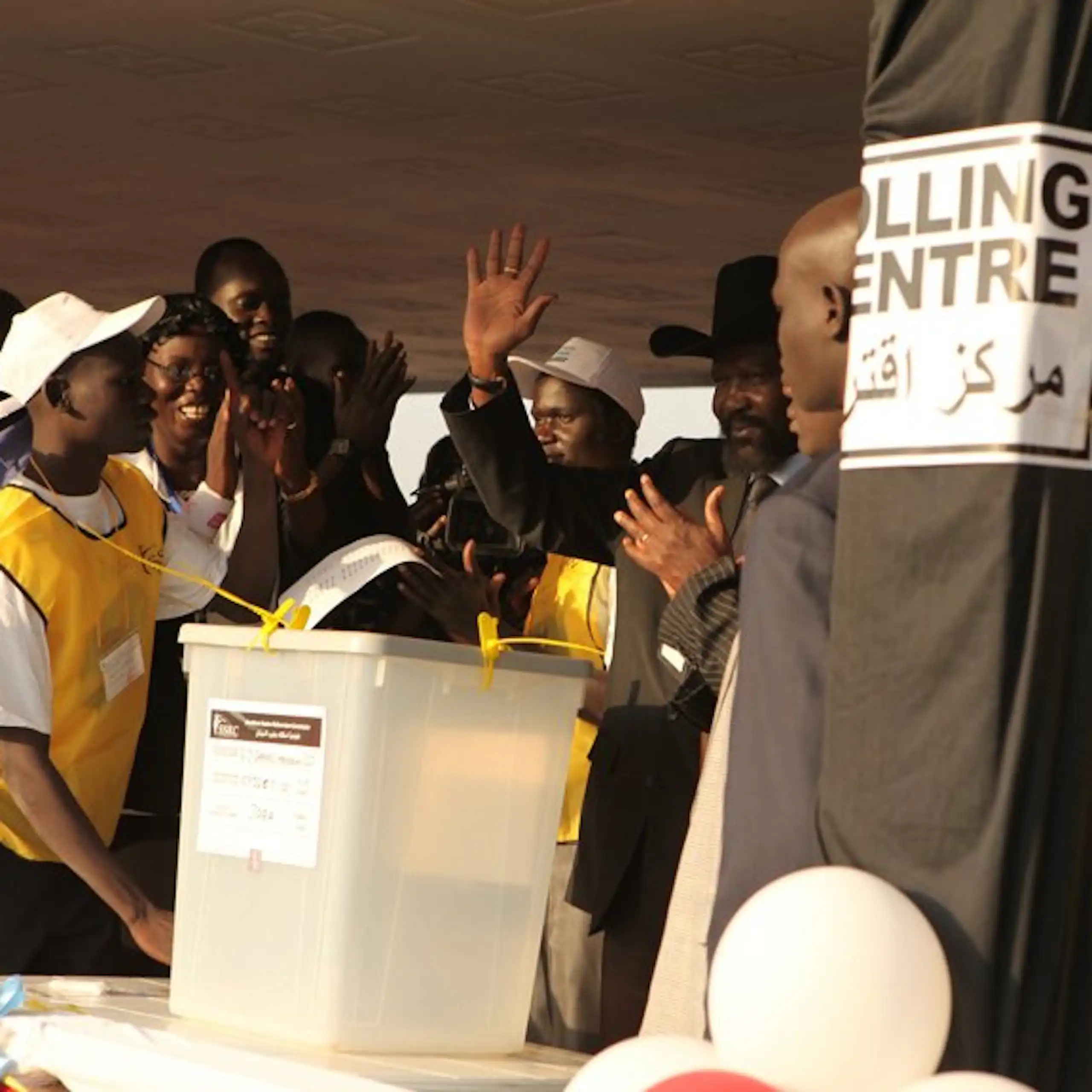 A man waving as he walks toward a transparent white box while surrounded by a small crowd of people