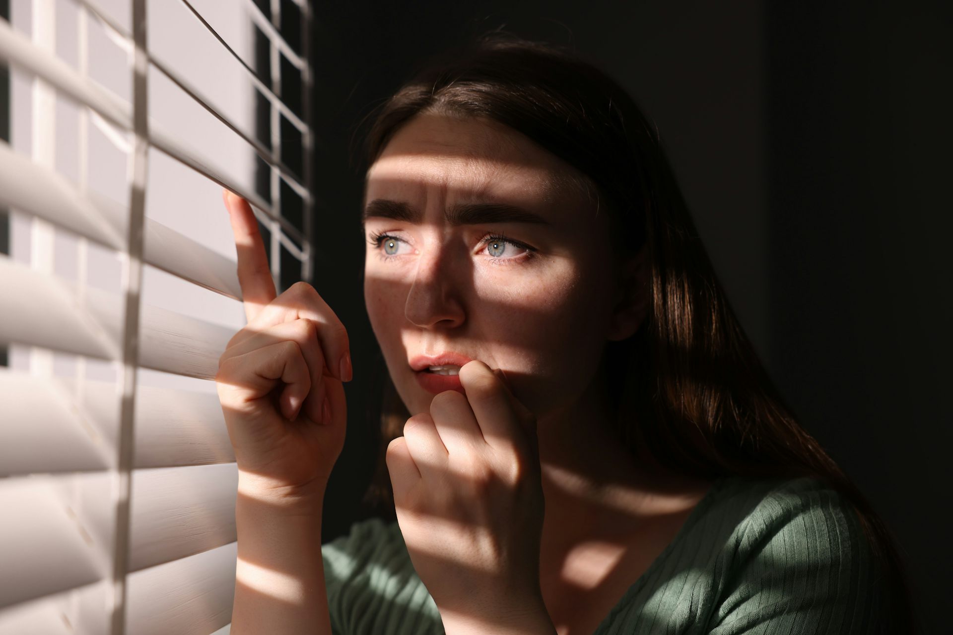 A woman looking fearfully out of window blinds