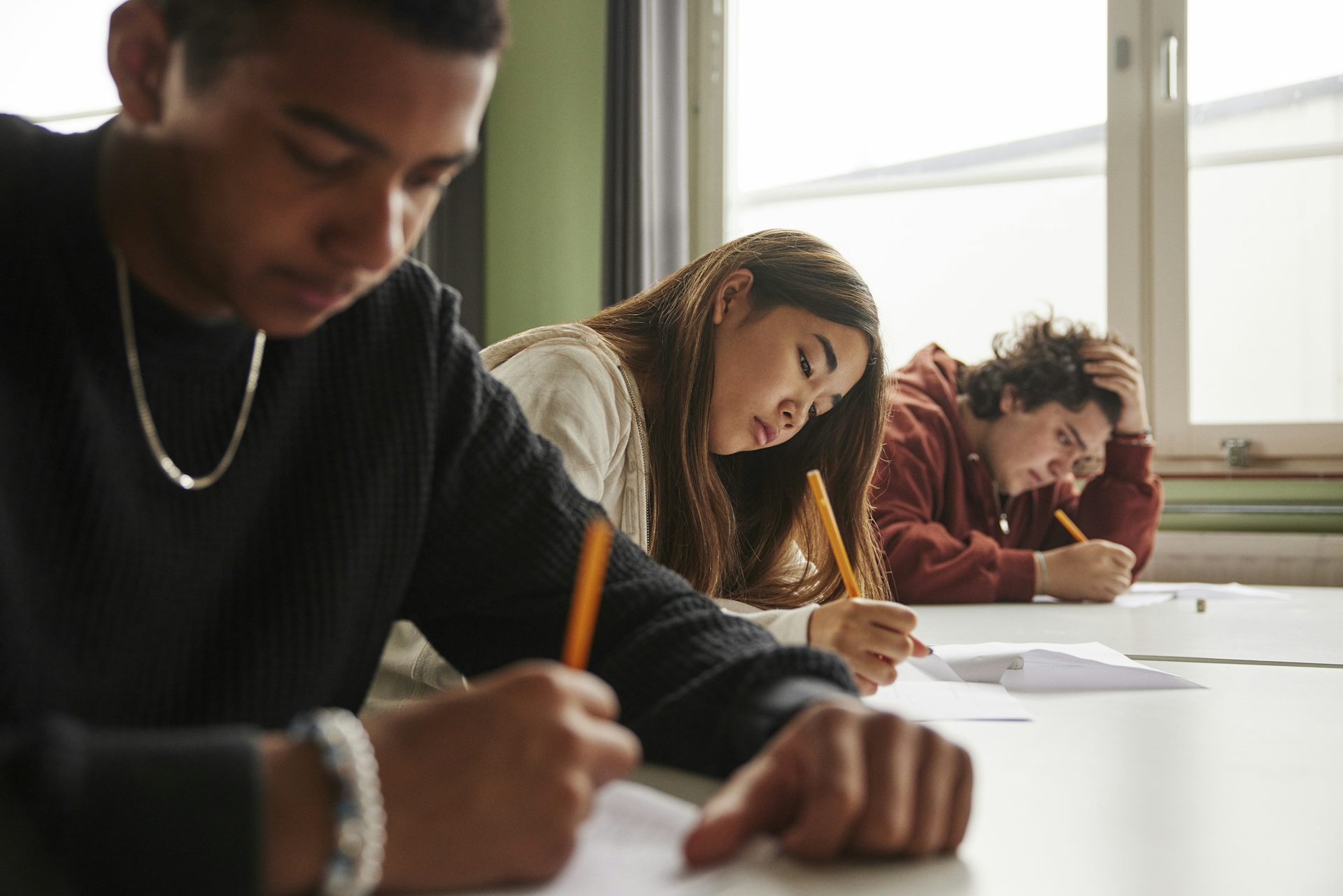 Three teenagers hold pencils and lean over a table. They sit in a row.
