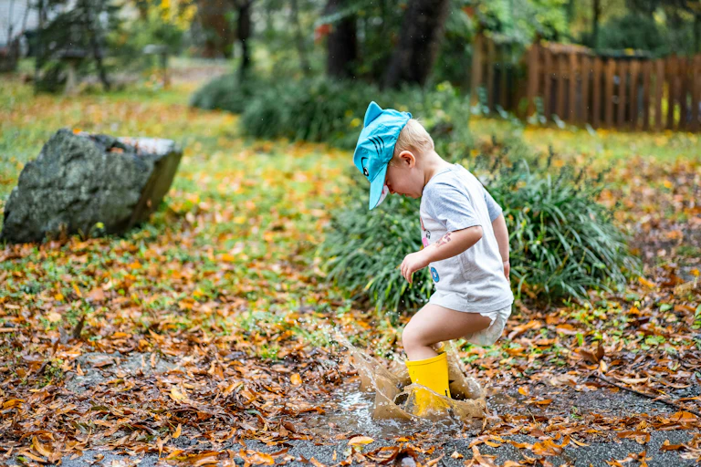 A toddler in a nappy, t-shirt and gumboots jumps in a puddle.