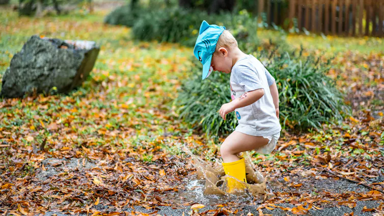 A toddler in a nappy, t-shirt and gumboots jumps in a puddle.