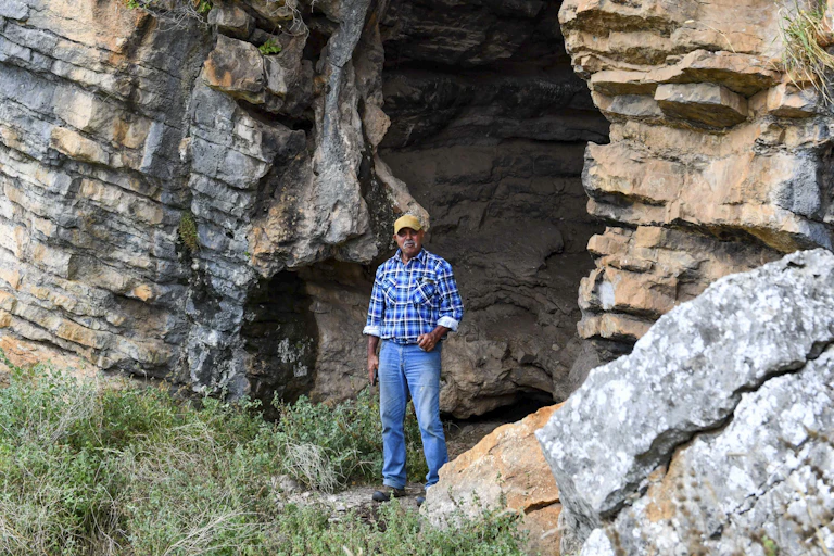A man in a blue shirt and jeans standing at the entrance of a large rocky cave.