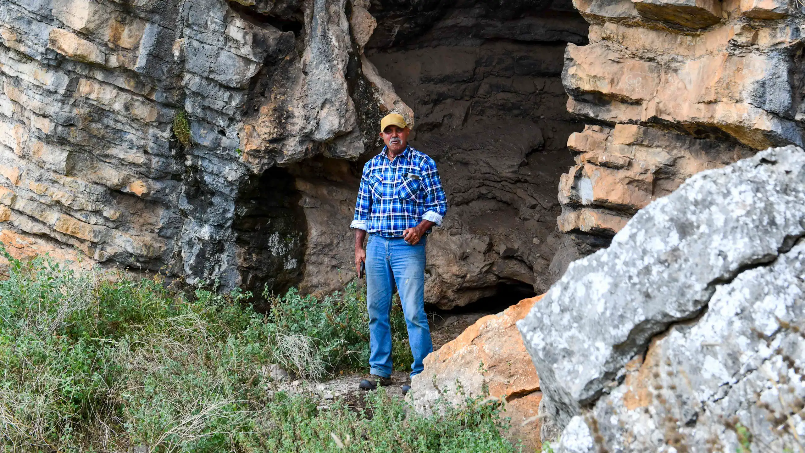A man in a blue shirt and jeans standing at the entrance of a large rocky cave.