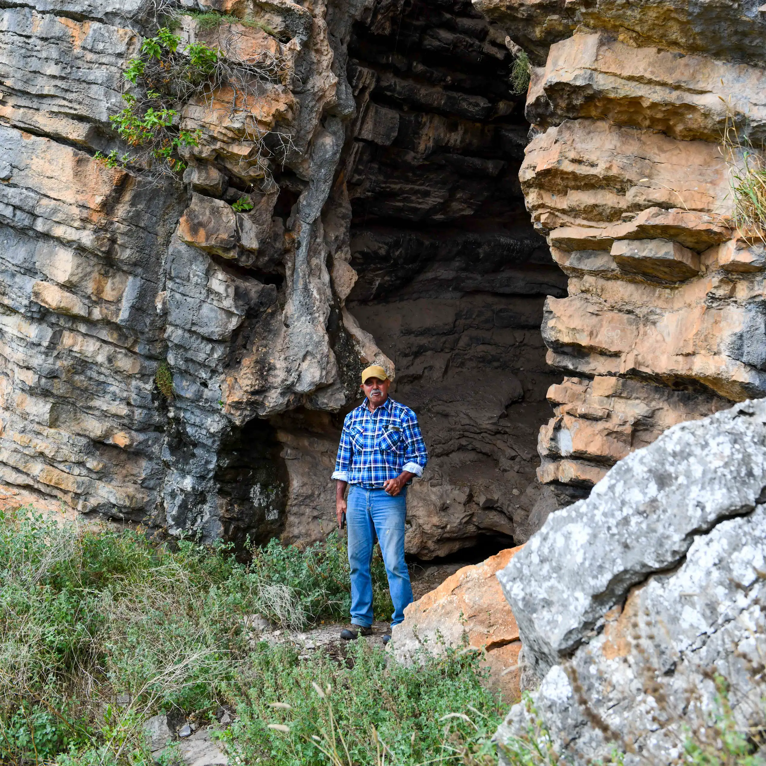 A man in a blue shirt and jeans standing at the entrance of a large rocky cave.