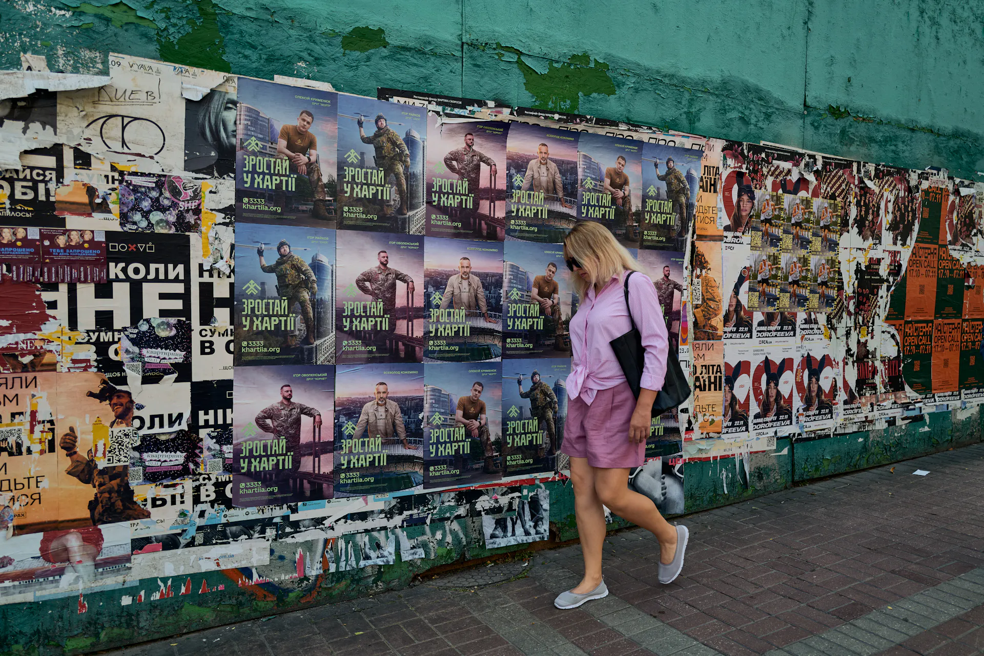 A woman walks down the street next to a wall with lots of posters.