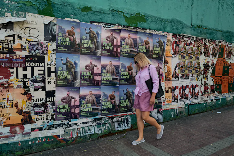 A woman walks down the street next to a wall with lots of posters.