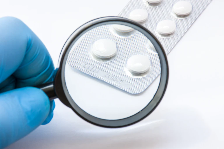 A gloved hand holding a magnifying glass over a batch code on a blisterpack of pills.