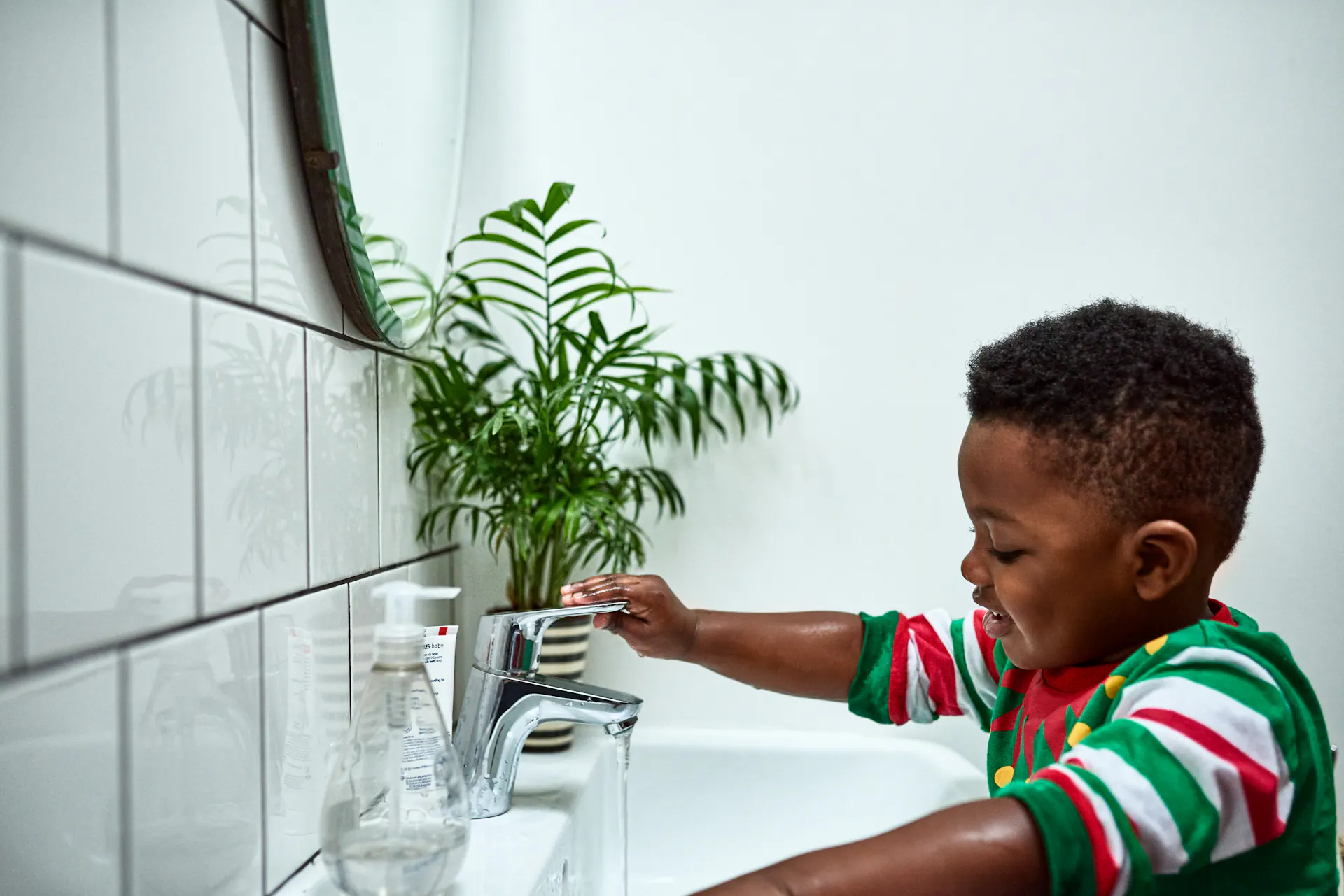 A young boy smiles and washes his hands at a white sink with a plant nearby.
