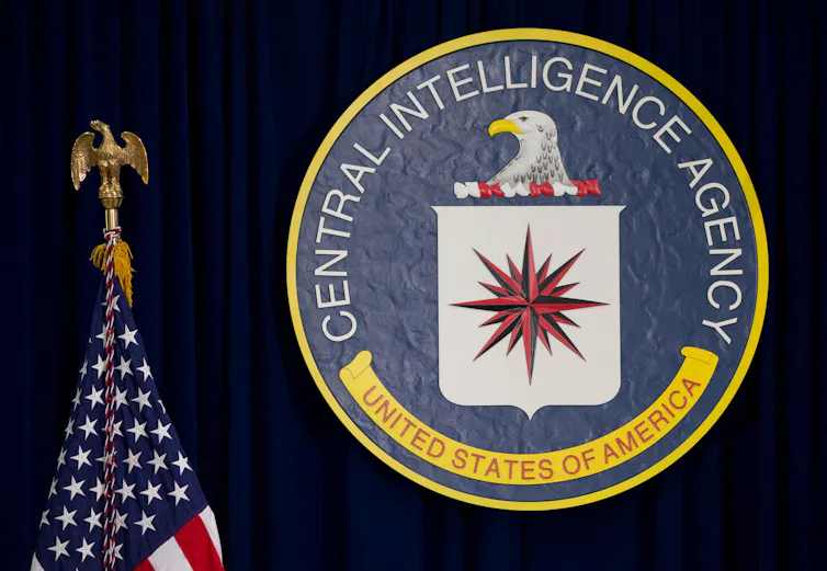 The round seal of the Central Intelligence Agency is in front of a dark blue curtain and beside a US flag