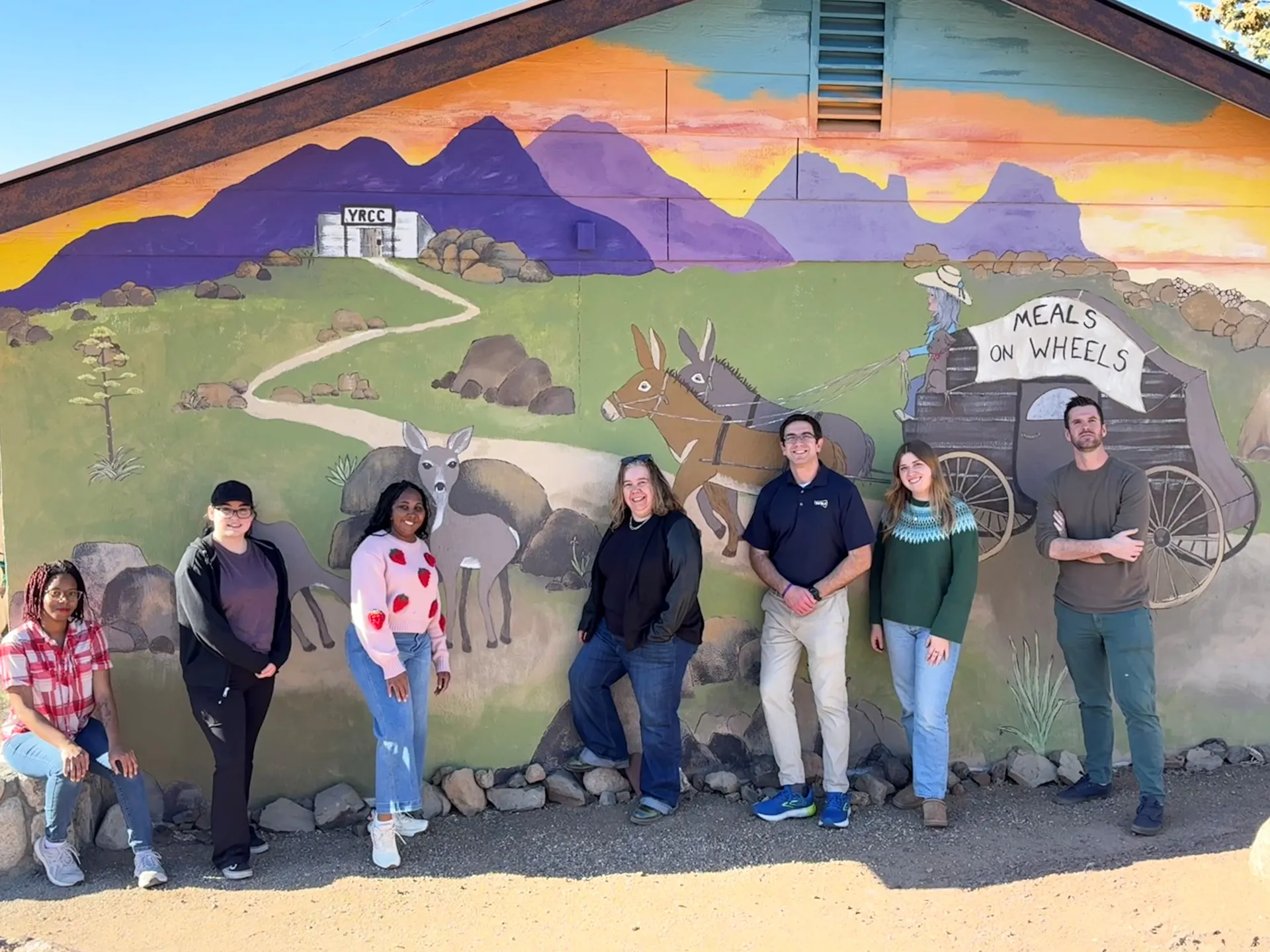Seven people stand in front of a colorful mural.