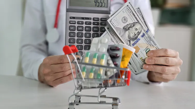 The headless torso of a doctor, wearing a stethoscope around their neck and holding a calculator in one hand and a stack of $100 bills, with a tiny shopping cart full of pills sitting in front of them