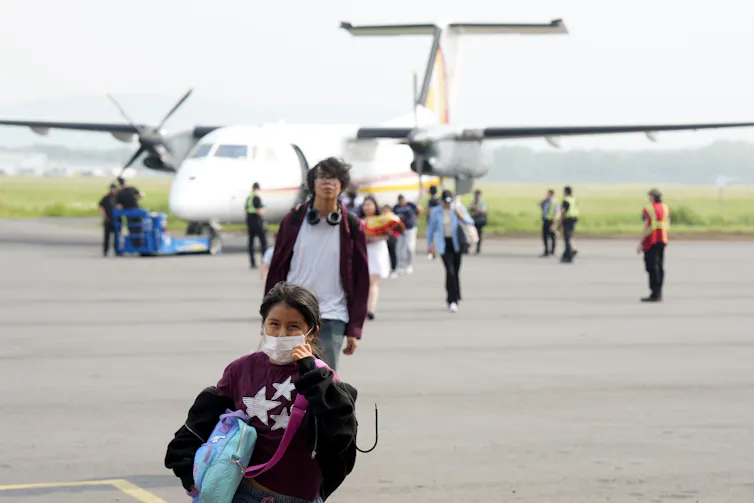 una niña con mascarilla y otras personas caminan desde el avión por la pista