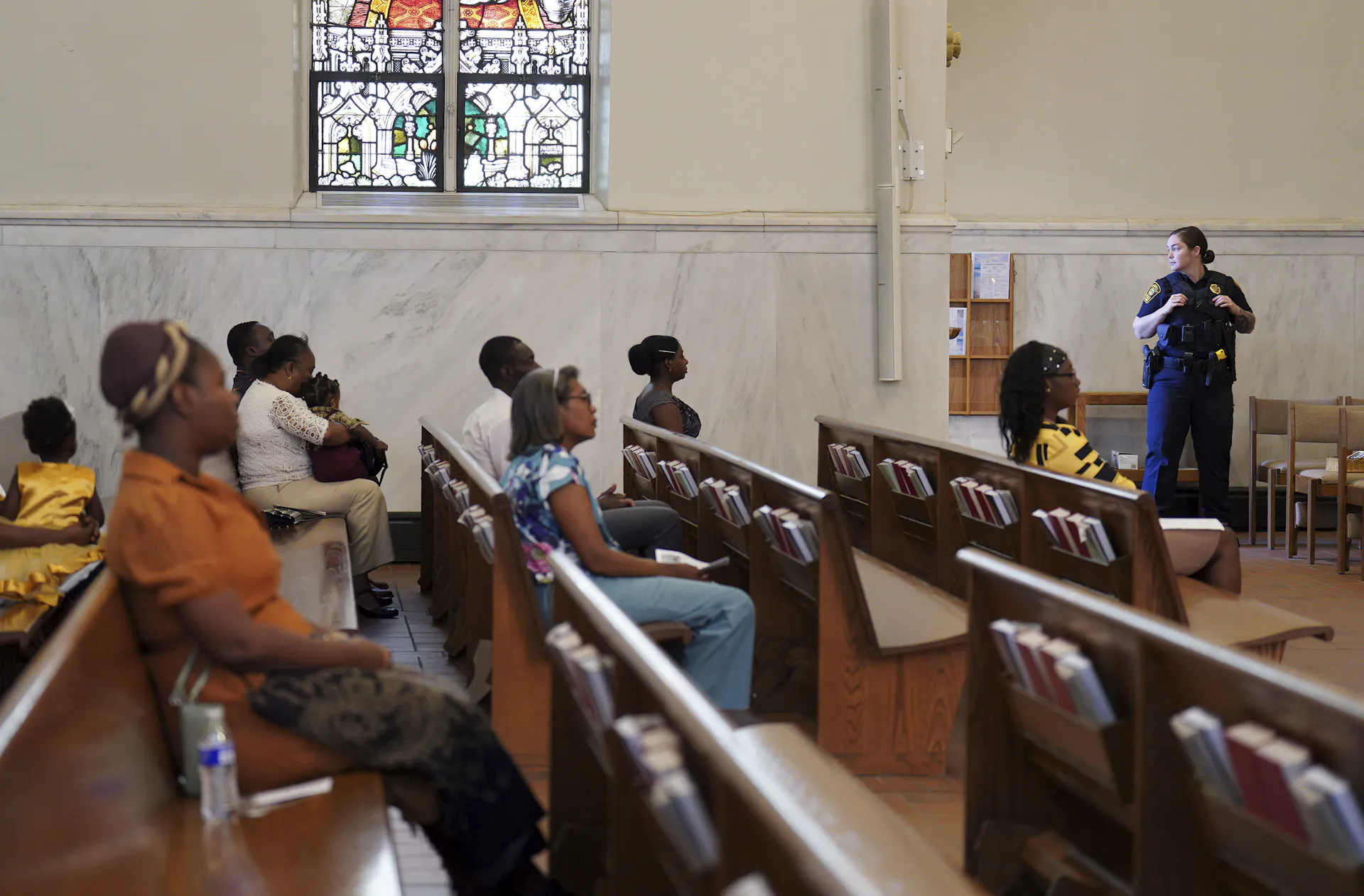 A woman in a blue uniform stands at the side of a sanctuary, in front of wooden pews with a few people seated in them.