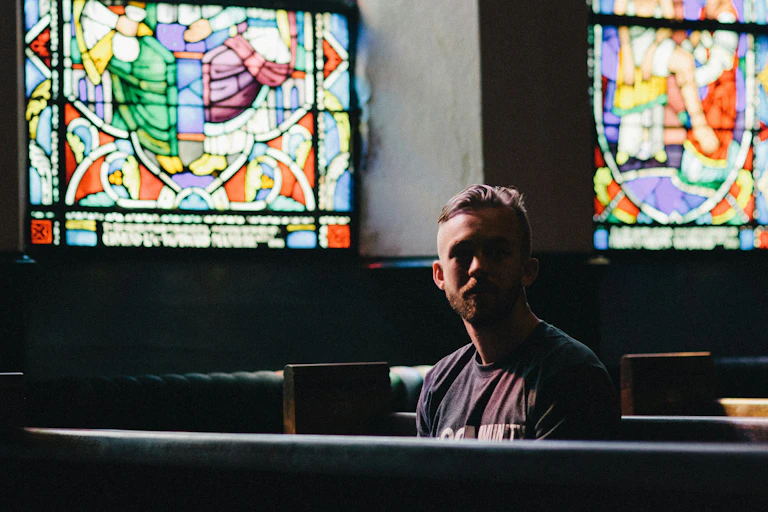 A young man sits on a church pew in front of stained glass windows