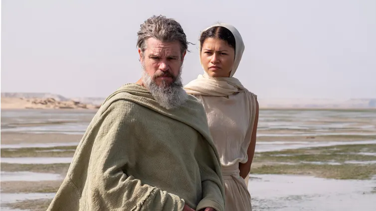 A man and woman stand in a deserted plain in ancient Greek-style clothing.